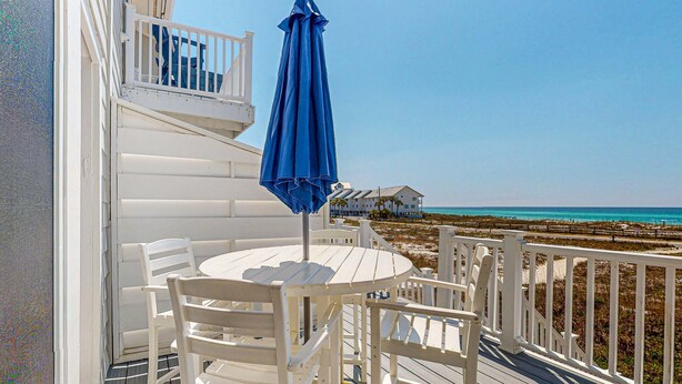 Eine helle Terrasse im Freien mit einem runden Tisch und Stühlen, einem blauen Sonnenschirm und Blick auf Strand und Meer.