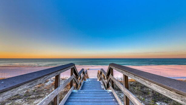 Eine Holzpromenade führt zu einem Strand mit blauem Himmel.