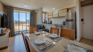A modern kitchen with a large refrigerator, a dining area with a table set for four, and a view of the city through large windows. - Mirante-506 - Apt 300m From the Beach by Carpediem (Fortaleza)