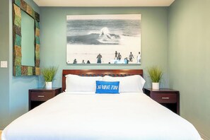 Quarto de hotel com uma cama, cabeceira, pintura pendurada na parede, duas mesinhas de cabeceira com plantas em vasos e um travesseiro com a inscrição 'NO WAKE ZONE'.