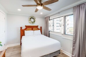A bedroom with a wooden bed, a ceiling fan, a window with curtains, and a view of buildings outside. - Beachfront condo in gated resort with pools, lake access, fast WiFi & patio (Panama City Beach)