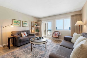 A living room with a sofa, armchair, coffee table, floor lamp, and a bookshelf. - Pinnacle Port getaway with balcony, pools & quick beach access (Panama City Beach)