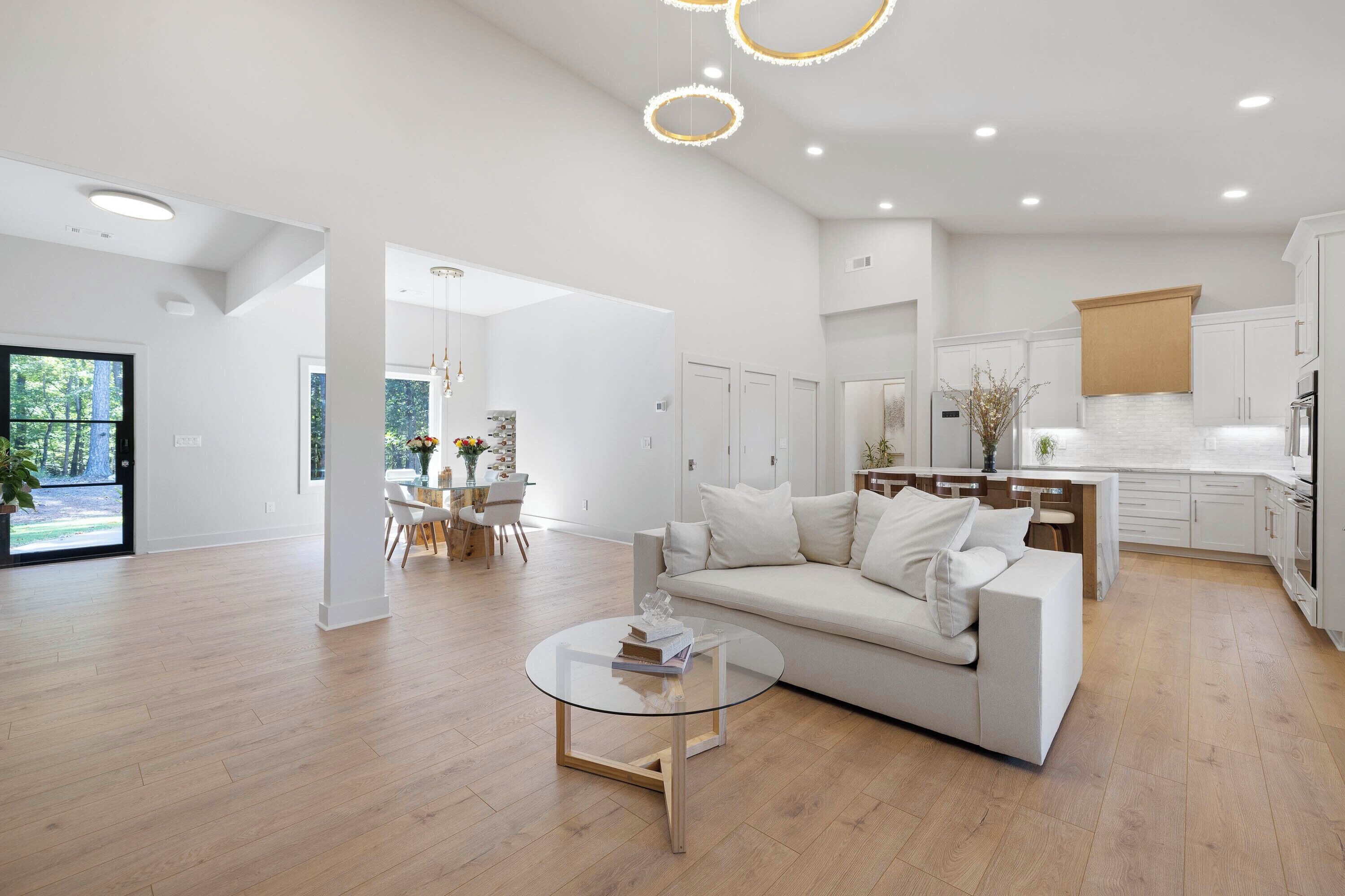 A modern living room with a white sofa, a glass coffee table, and a kitchen area with white cabinets and a wooden countertop.