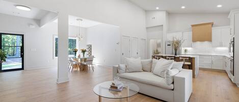 A modern living room with a white sofa, a glass coffee table, and a kitchen area with white cabinets and a wooden countertop.