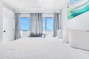 Ein Schlafzimmer mit einem großen Bett, grauen Vorhängen, Blick zum Himmel durch das Fenster und einem Gemälde an der Wand.