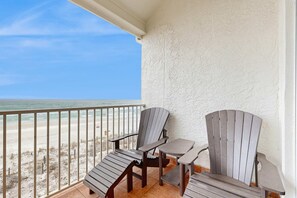 Un balcon avec deux fauteuils Adirondack et une petite table, donnant sur une plage bordée d’une clôture et de dunes de sable.