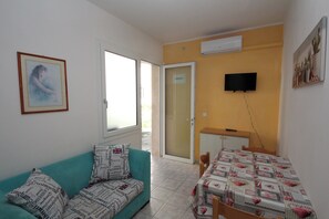 A small, well-lit room with a teal sofa, a dining table with a patterned tablecloth, a television mounted on the wall, and a door leading outside. - Rosolina Mare Flat Near Beach (Rosolina)