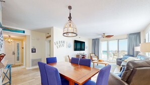 Una sala de estar moderna con una mesa de comedor de madera, sillas azules, un televisor grande y un balcón con vistas.