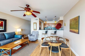 Un salon moderne avec un canapé, une table basse et un coin repas comprenant des chaises et une table en verre. On peut voir un ventilateur de plafond et des luminaires suspendus.