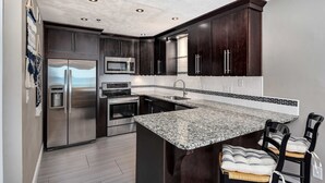A modern kitchen with dark wood cabinets, stainless steel appliances, a granite countertop, and a built-in island with bar stools. - Stylish oceanfront condo w/ pool access, private W/D, ocean views, & central AC (Santa Rosa Beach)