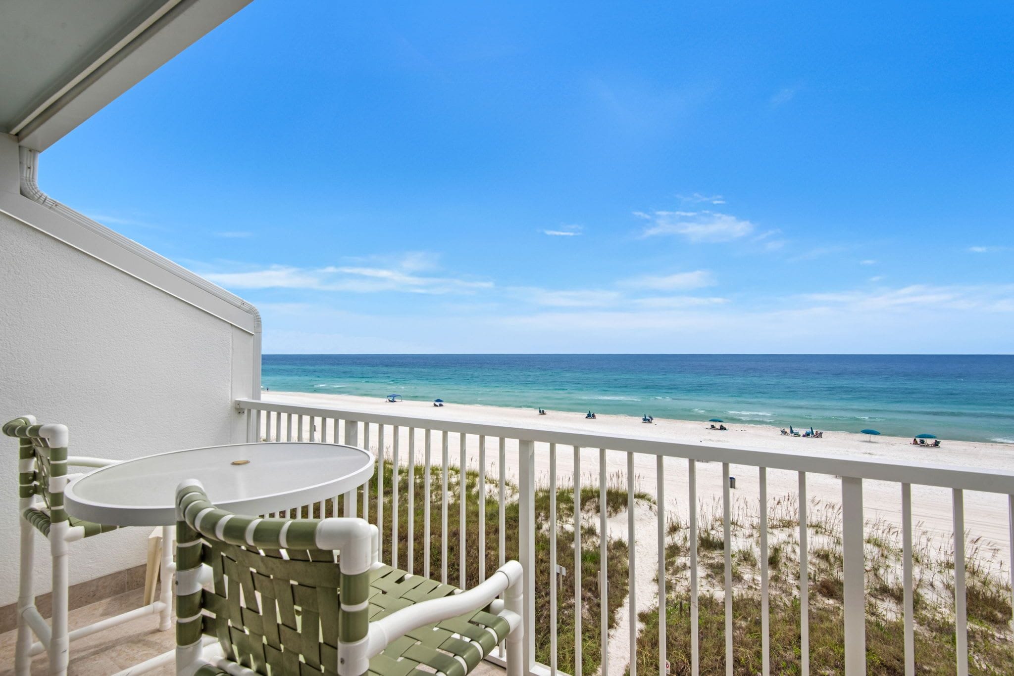 A balcony with a table and chairs overlooking a beach with people and umbrellas.