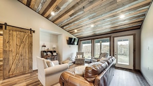 Ein geräumiges Wohnzimmer mit Holzbalkendecke, einem großen Ledersofa, einem weißen Sessel und einem an der Wand befestigten Fernseher.
