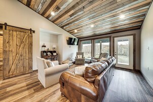 Un salón amplio con techo de madera, un sofá grande de cuero, un sillón blanco y un televisor montado en la pared.