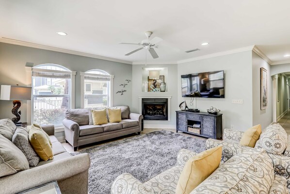 A living room with a grey sofa, a patterned sofa, and a grey armchair. There is a fireplace, a ceiling fan, and a TV mounted on the wall. - Coastal chic condo steps from the sand, shared pool & central location (Santa Rosa Beach)