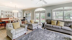 Un vaste salon doté d’un canapé gris, de fauteuils à motifs, d’un coin repas avec une table et des chaises en bois, et de grandes fenêtres offrant une vue sur l’extérieur.