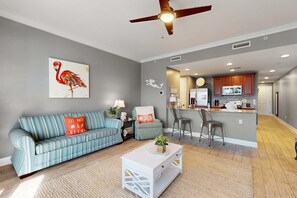 A living room with a sofa, a coffee table, and a painting of a flamingo. There is a kitchen area with a bar and a refrigerator. - 3rd-floor beach view condo with Pool, Hot tub, & private balcony (Panama City Beach)