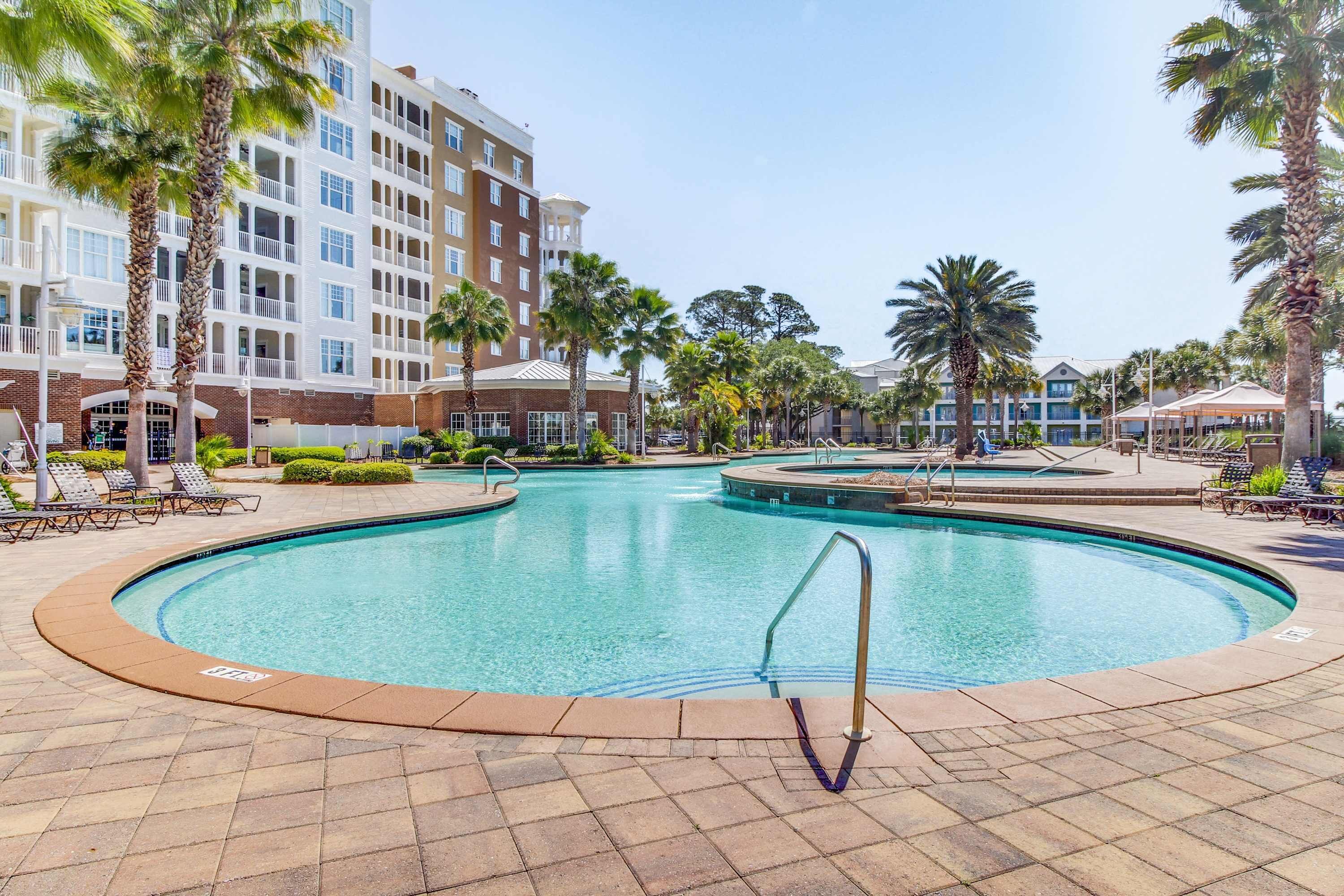 A swimming pool with a tiled deck, surrounded by palm trees and a multi-story hotel building.