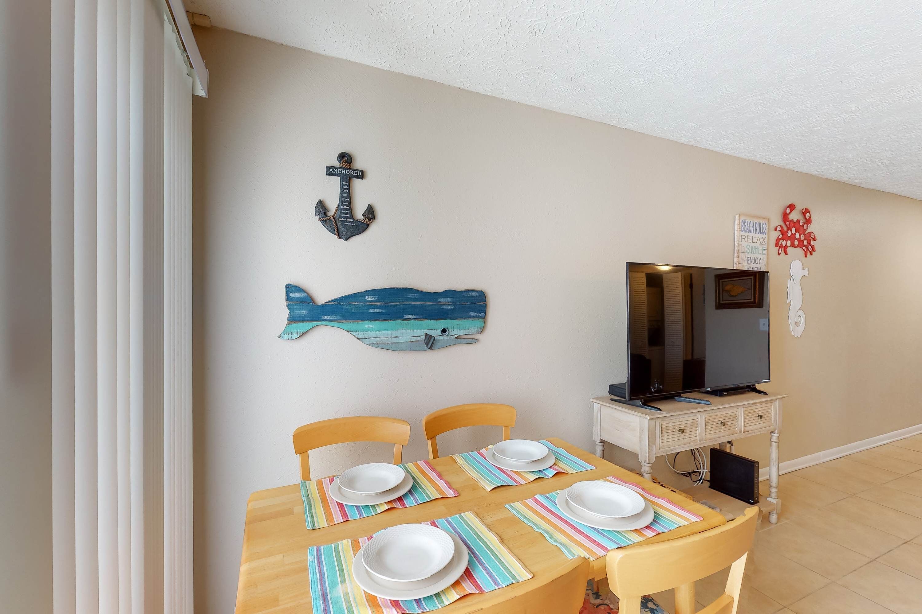 A dining area with a wooden table set for four, a television on a stand, and nautical-themed wall decorations.