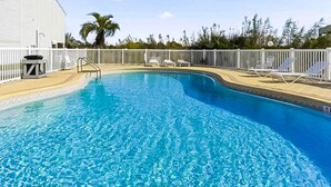 Une piscine entourée d’une clôture blanche, avec des chaises longues et un bâtiment visible au loin.