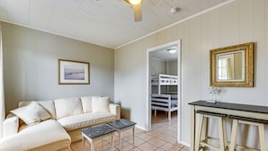 A room with a white sofa, two small tables, a wooden bar stool, a framed picture on the wall, and a bunk bed in the background.