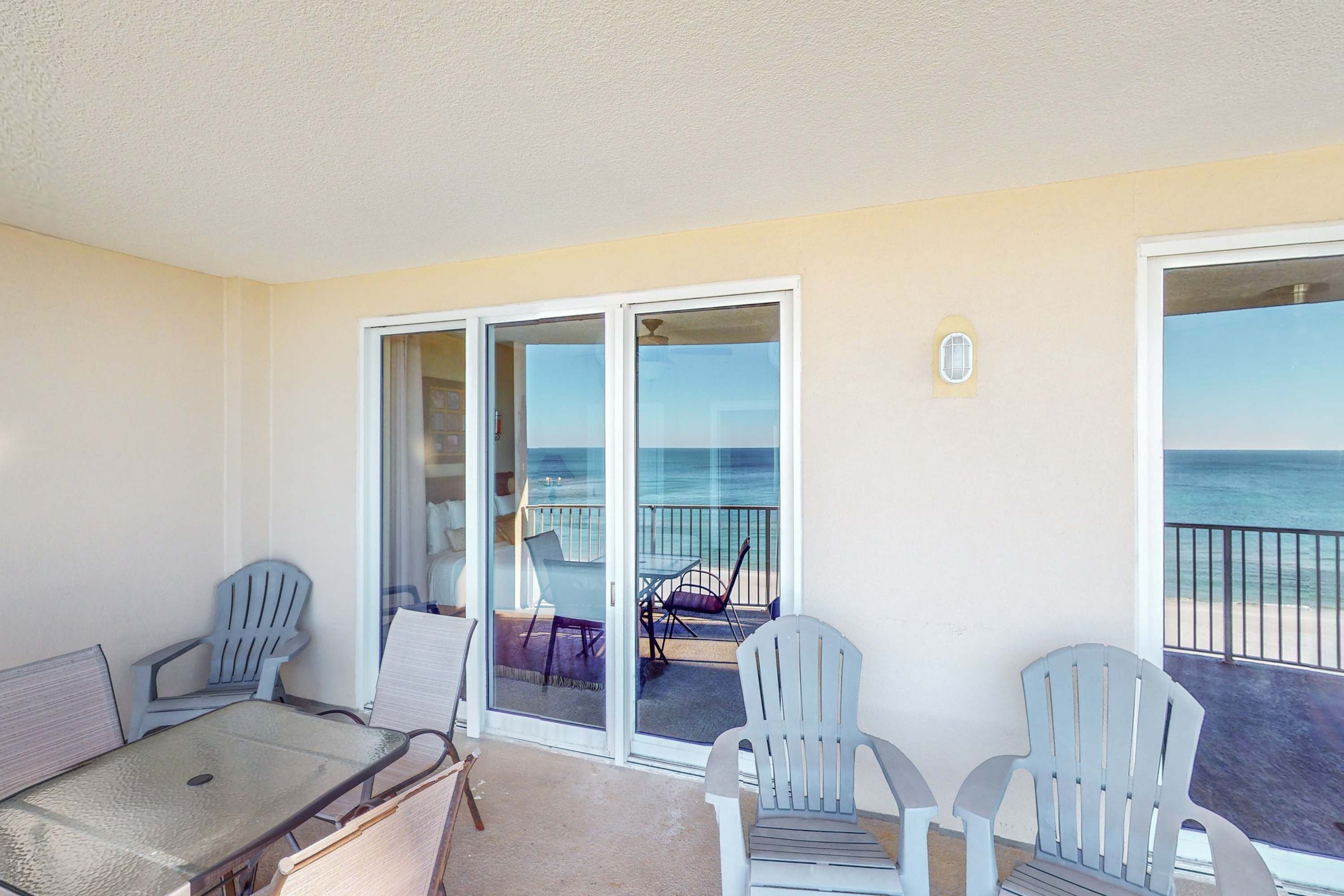 A balcony with a table and chairs, a sliding glass door leading to a beach view, and a small outdoor light fixture.