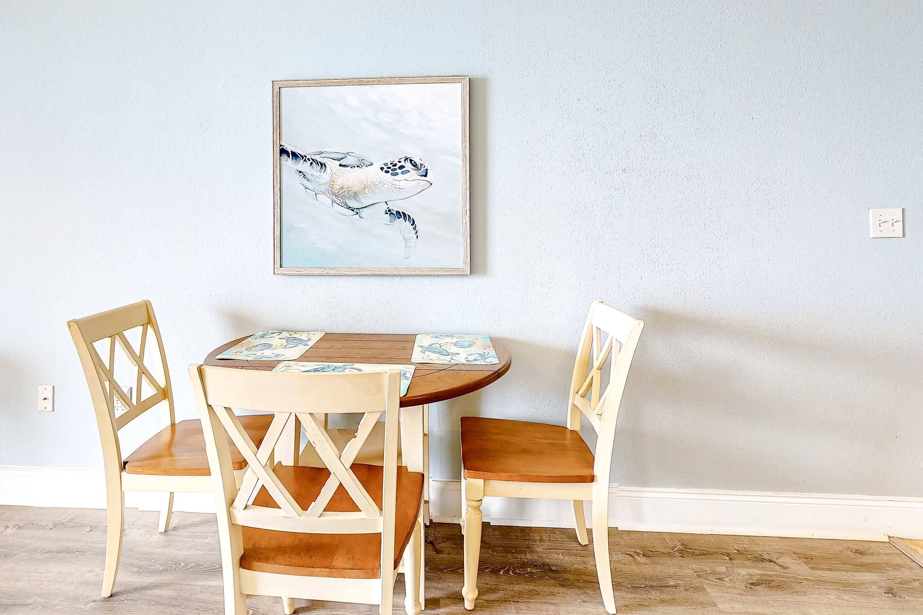 A dining area with a round table, two wooden chairs, and a framed artwork of a sea turtle on the wall.