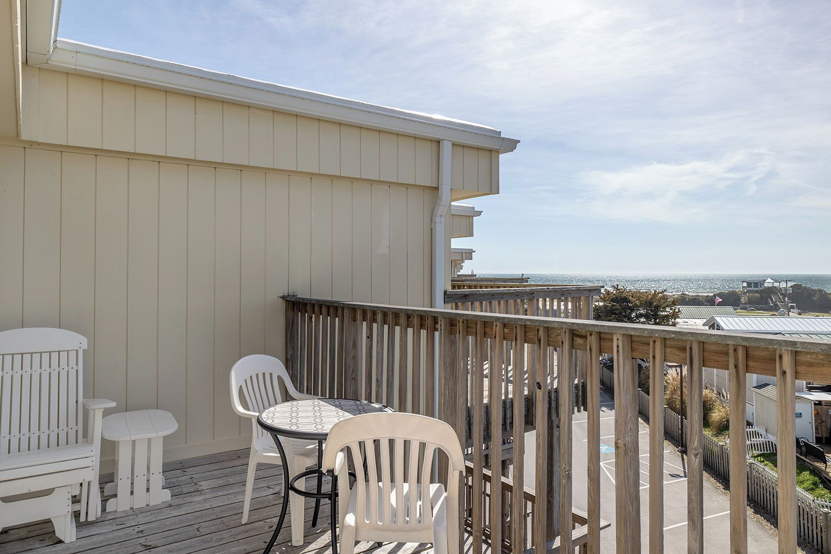 A balcony with a view of the ocean, white outdoor furniture, and a wooden railing.