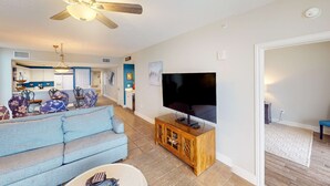 Un salon avec un canapé, un téléviseur, un meuble en bois, un ventilateur de plafond et, au fond, un coin cuisine.