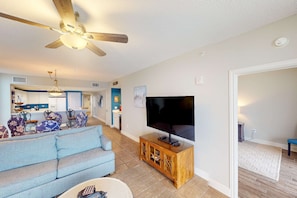 A living room with a sofa, TV, wooden cabinet, ceiling fan, and a kitchen area in the background. - Gulf-front condo with 4 pools, movie theatre, game room, 3 hot tubs, tennis, W/D (Panama City Beach)