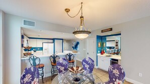 Un coin repas avec une table ronde en verre et des chaises à motifs floraux violets, un lustre et une cuisine en arrière-plan.