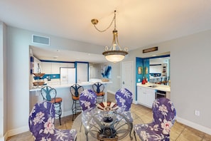 A dining area with a round glass table and purple floral chairs, a chandelier, and a kitchen in the background. - Gulf-front condo with 4 pools, movie theatre, game room, 3 hot tubs, tennis, W/D (Panama City Beach)