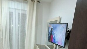 A bedroom with a bed, a TV mounted on the wall, a small white cabinet, and a wooden door. - F2 high standings (Béjaïa)