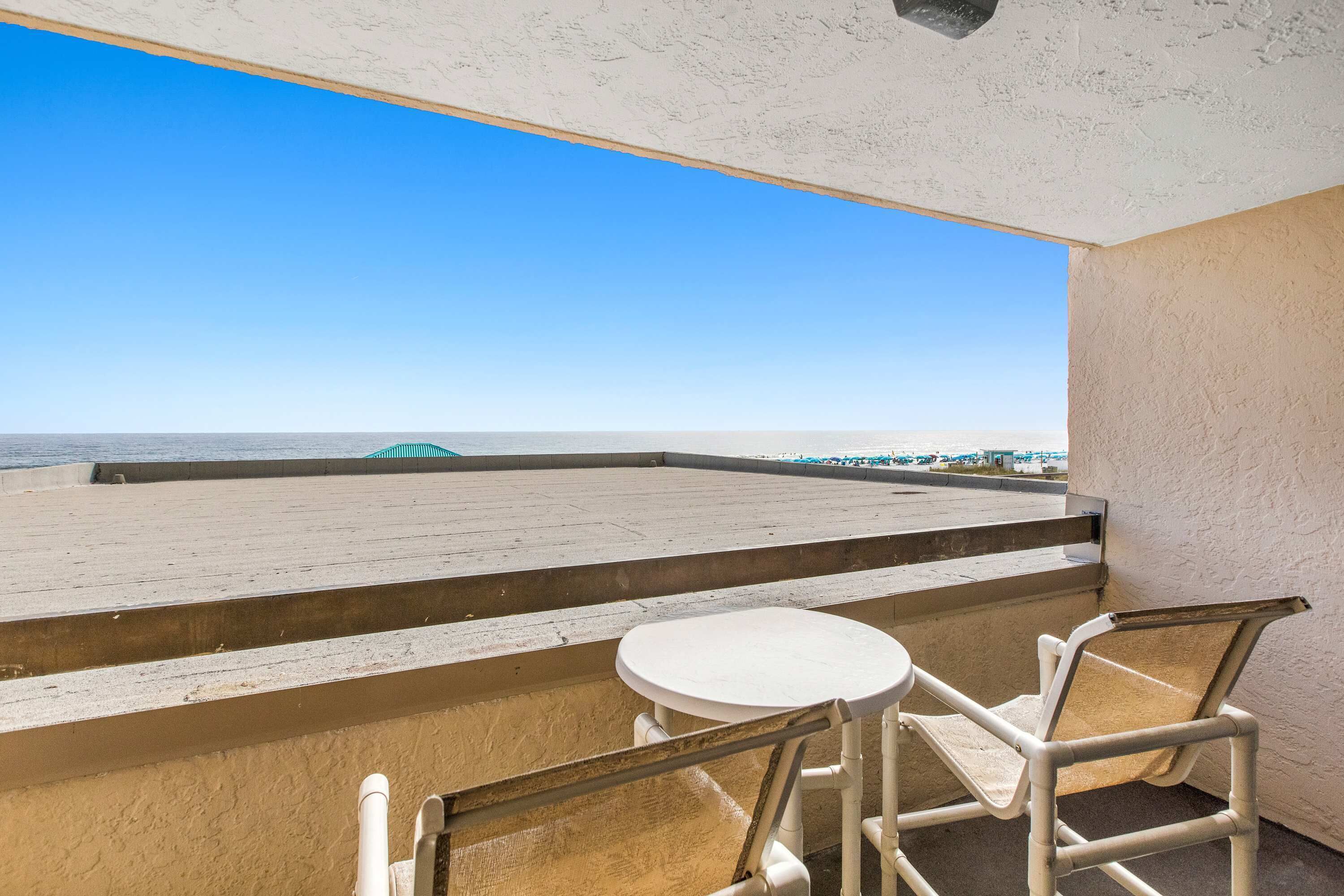 A balcony with a white table and chairs, overlooking a beach with a clear sky.