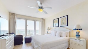 Quarto de hotel com cama, mesa de cabeceira, cômoda, ventilador de teto e uma grande janela com cortinas.