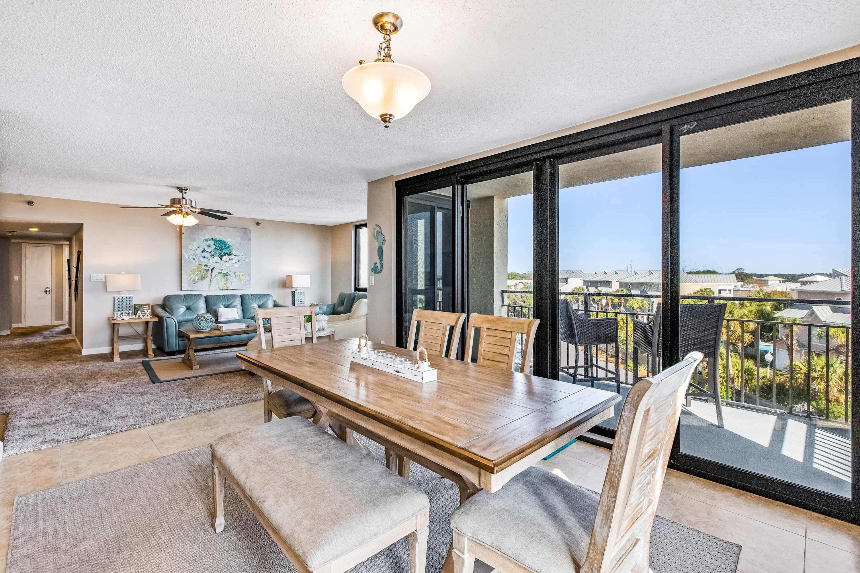 A spacious living area with a large wooden dining table, comfortable seating, a ceiling fan, and a balcony with a view.