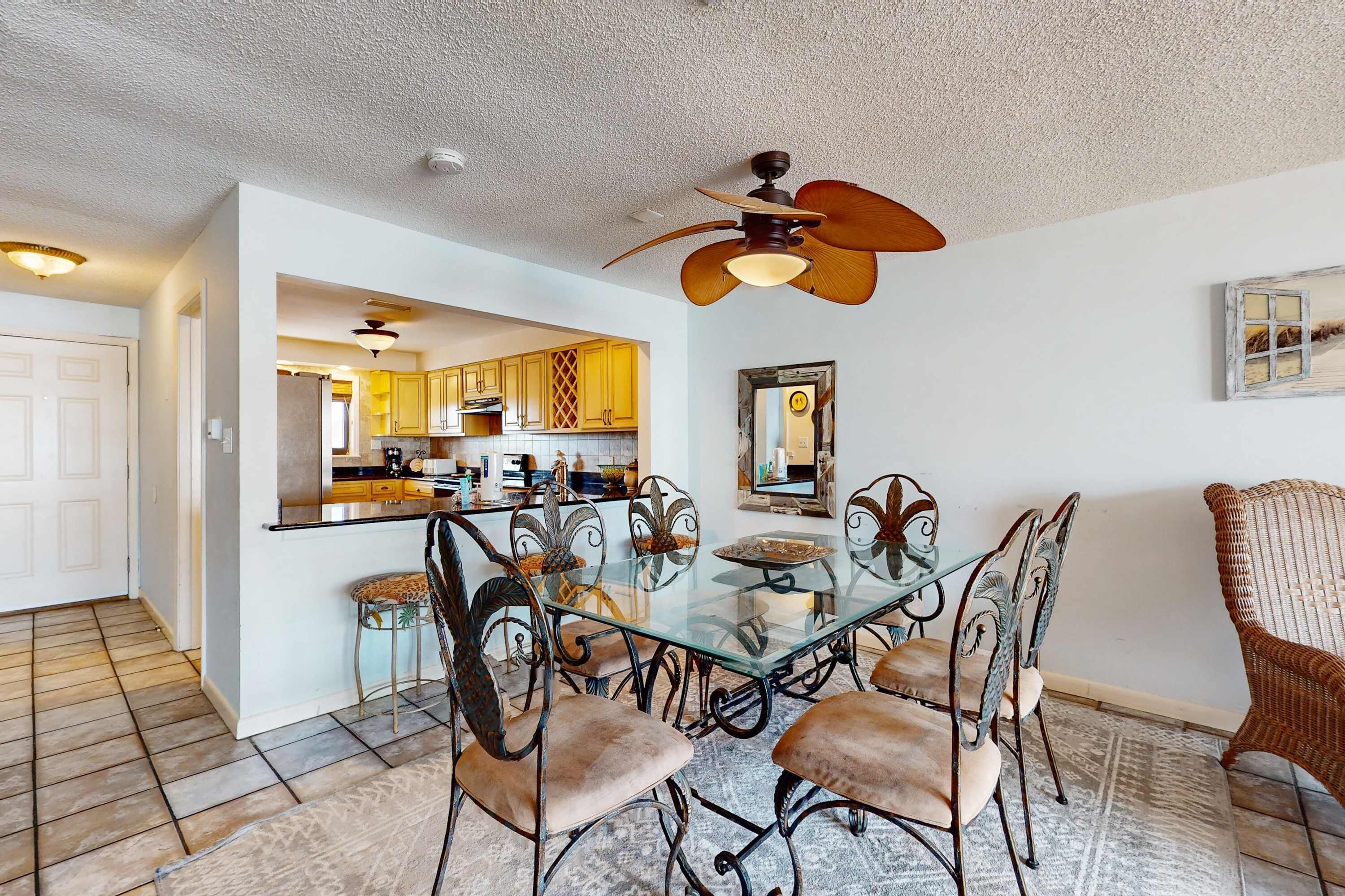 A dining area with a glass table and metal chairs, a ceiling fan, and a kitchen visible through an open doorway.