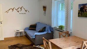 A cozy living room with a sofa, a small round table, a wooden dining table, a cowhide rug, a wall-mounted plant, and a mountain graphic.