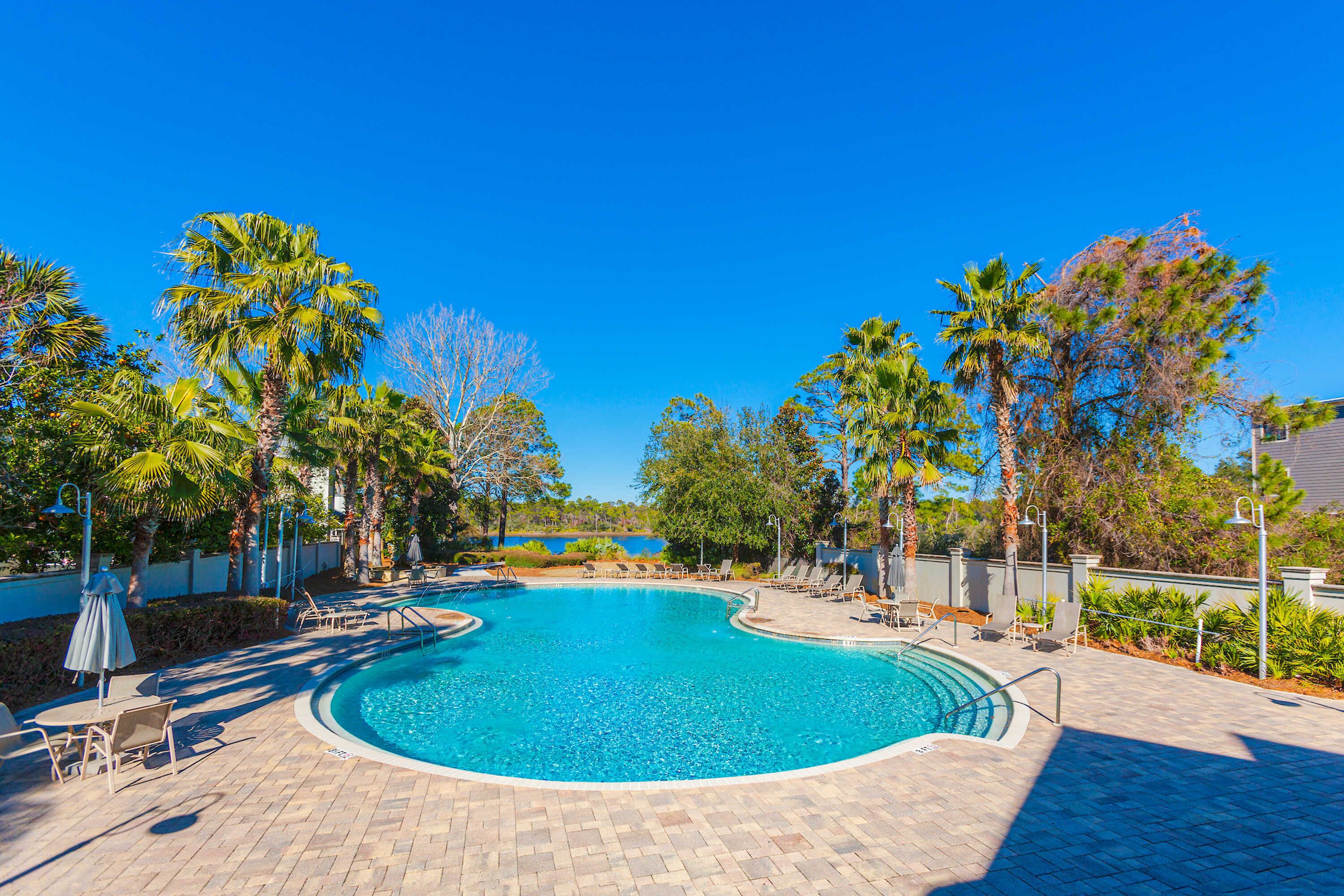 Una piscina azul cristalina rodeada de sillas de descanso y sombrillas, con palmeras y una vista del agua en el fondo.