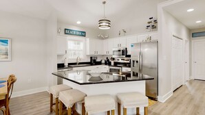 A modern kitchen with white cabinetry, a large island with stools, stainless steel appliances, and a 'Beach House' sign. - Dog-friendly home with pool, patio, & washer&dryer (Santa Rosa Beach)
