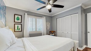 Quarto com cama, ventoinha no teto, janela com persianas, guarda-roupa e prateleira na parede.