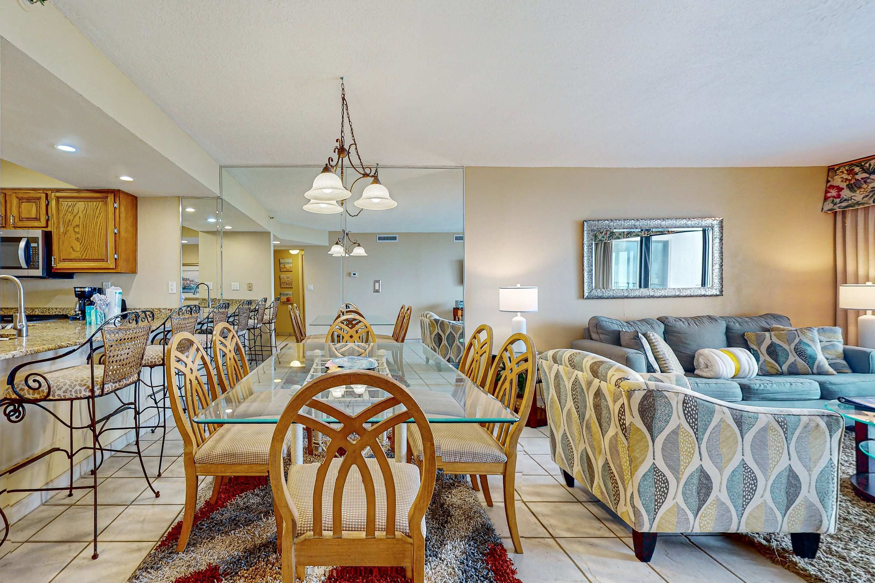 Un vaste espace de vie comprenant une table à manger avec un plateau en verre, entourée de chaises en bois, d’un canapé à motifs et d’un grand miroir mural.