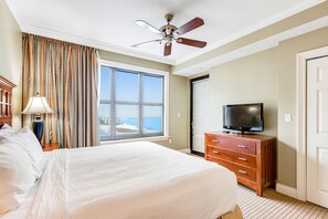 Ein Hotelzimmer mit einem großen Bett, einem Deckenventilator, einem Fernseher auf einer Holzkommode und Meerblick durch ein großes Fenster.
