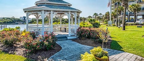 A gazebo with a white roof and open sides, surrounded by a well-maintained garden with various plants and flowers.