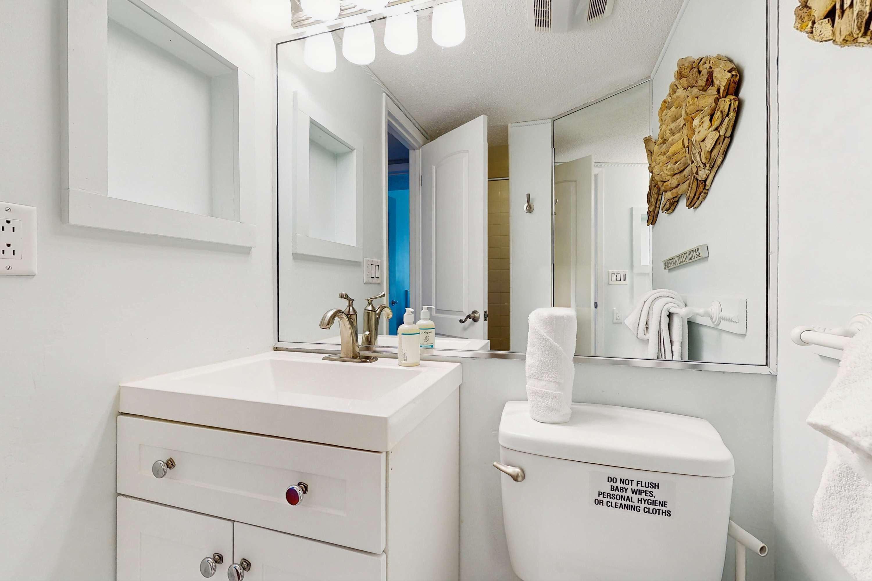 A modern bathroom with a white sink, mirror, and toilet. There is a towel rack on the wall and a decorative item above the mirror.