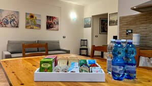 Uma área de refeições com uma mesa de madeira, um sofá cinza e uma cadeira. Há garrafas de água e uma bandeja com alimentos na mesa.
