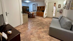 Uma sala de estar com sofá cinzento, uma área de jantar com mobiliário de madeira e uma cozinha com armários de madeira e forno embutido.