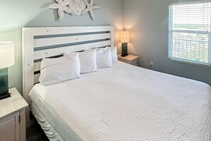 Un lit bien fait, avec du linge de lit et des oreillers blancs, une étoile de mer décorative accrochée au mur, une lampe sur une table de chevet et une fenêtre avec des stores.
