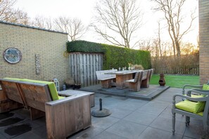 An outdoor patio with wooden furniture, a brick wall, and a hedge. - 3-bedroom House Near North Sea (Sint Maartensvlotbrug)