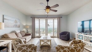 Una sala de estar con ventilador de techo, televisor, sofá, dos sillones, una mesa de centro y un amplio ventanal con cortinas.