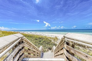 Eine Holztreppe führt zu einem Strand mit blauem Himmel und ruhigem Wasser.